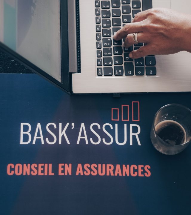 Main qui tape sur un ordinateur avec une tasse de café et le logo de Bask'Assur