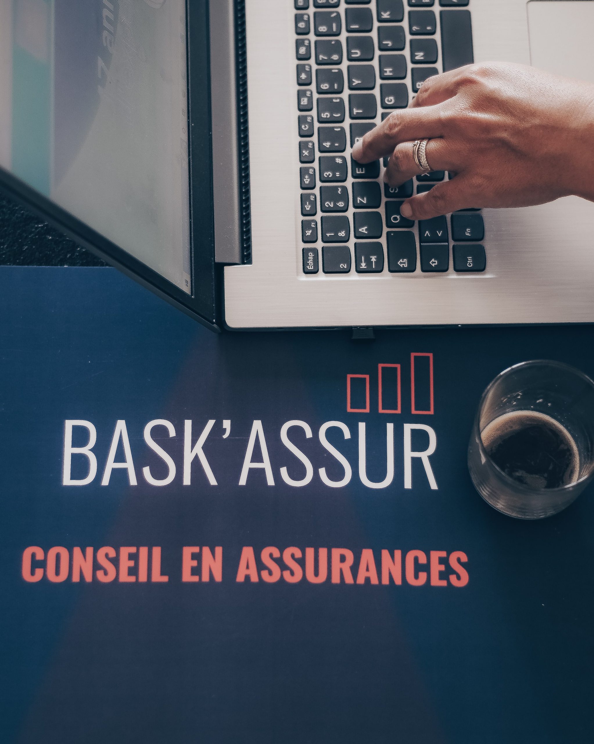Main qui tape sur un ordinateur avec une tasse de café et le logo de Bask'Assur
